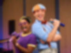 Blippi and Meekah happily sing together with microphones onstage.